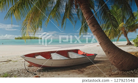 Small red and white boat resting on a pristine white sand beach under the shade of palm trees in Aitutaki, Cook Islands, with turquoise waters and a small island in the background Small red and white boat resting on a pristine white sand beach under the shade of palm trees in Aitutaki, Cook Islands, with turquoise waters and a small island in the background 127925548