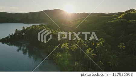 Aerial drone footage revealing the stunning beauty of a tropical island coastline at sunrise, with swaying palm trees and rolling hills glowing in golden light. Amazing nature travel background 127925568