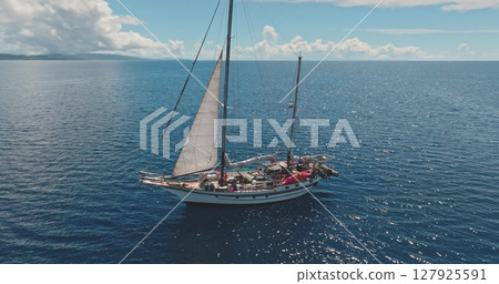 Aerial drone footage of a ketch sailing near a tropical island on a sunny day, with white sails and a wooden deck, surrounded by blue waters and a distant coastline. Wild sea nature travel background 127925591