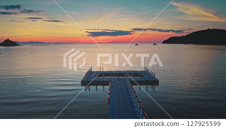 Plastic floating dock extends into calm ocean water at sunset with vibrant colorful sky, distant islands, and boats creating a serene and picturesque scene Plastic floating dock extends into calm ocean water at sunset with vibrant colorful sky, distant islands, and boats creating a serene and picturesque scene 127925599