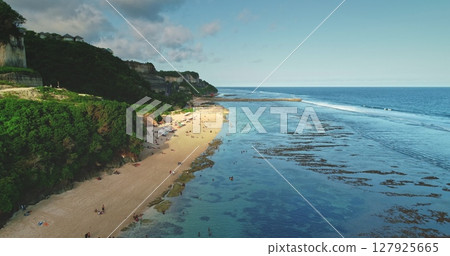 Crystal-clear turquoise water lapping against a sandy shore at a picturesque Bali beach, framed by lush cliffs and bustling tourists enjoying the tropical paradise Crystal-clear turquoise water lapping against a sandy shore at a picturesque Bali beach, framed by lush cliffs and bustling tourists enjoying the tropical paradise 127925665