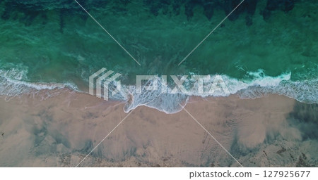 Aerial drone view of turquoise ocean waves crashing on the sandy shore of Bias Tugel Beach in Bali, Indonesia, creating a beautiful contrast between the vibrant water and the light-colored sand 127925677