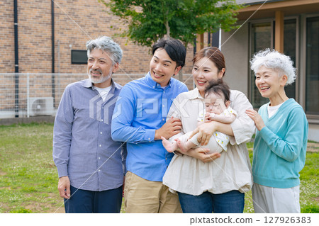 A three-generation family smiling and surrounding a baby in front of their house 127926383