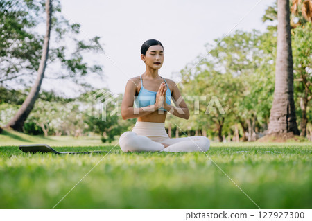Asian woman meditating in a peaceful park setting, sitting cross-legged on green grass 127927300