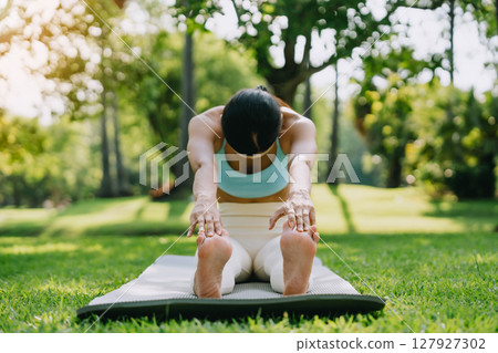 Young Asian woman doing yoga stretching on green grass with trees in the background on a sunny day 127927302