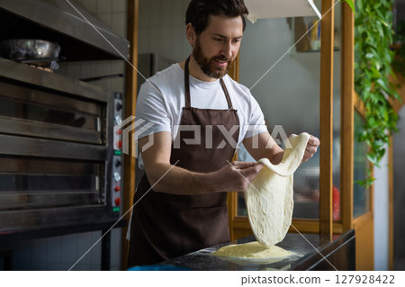 Bearded man wearing a white t-shirt and brown apron tosses pizza dough in a bakery kitchen Bearded man wearing a white t-shirt and brown apron tosses pizza dough in a bakery kitchen 127928422