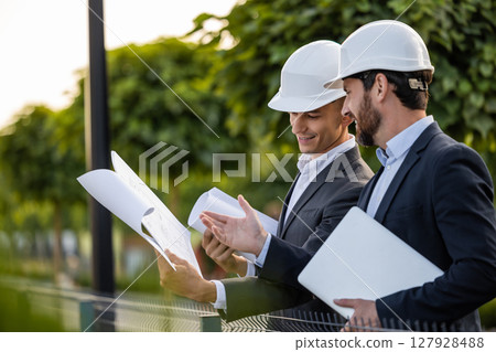 Engineers reviewing blueprints with laptop outdoors 127928488