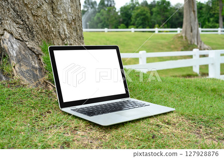 Laptop with blank screen laptop on fresh green grass near a tree trunk 127928876