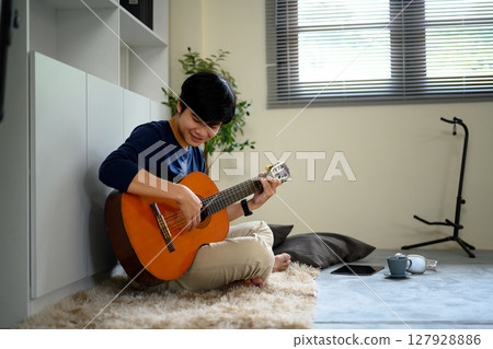 Happy asian in comfortable clothes playing guitar on the floor in a living room Happy asian in comfortable clothes playing guitar on the floor in a living room 127928886