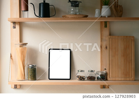 Digital tablet with blank screen on kitchen shelf surrounded by glass jars, pasta, and spices 127928903