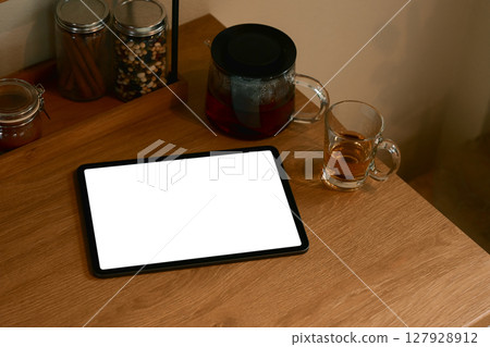 Digital tablet with blank screen on kitchen table beside glass teapot and cup of tea Digital tablet with blank screen on kitchen table beside glass teapot and cup of tea 127928912
