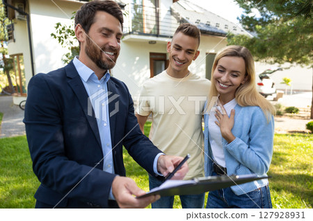 Real estate agent presenting clipboard with documents to couple in front of residential property 127928931