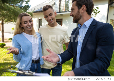 Real estate agent discussing property details with couple using clipboard in front of house 127928934