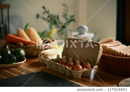 Fresh organic eggs in cardboard carton on a wooden kitchen counter surrounded by vegetables 127928943
