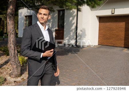 Real estate agent posing with clipboard and hand in pocket in front of house with garage Real estate agent posing with clipboard and hand in pocket in front of house with garage 127928946