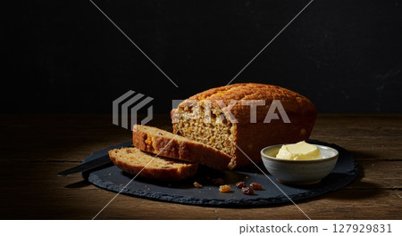 Homemade fruit loaf cake with raisins and butter served on a rustic slate board with low light Homemade fruit loaf cake with raisins and butter served on a rustic slate board with low light 127929831