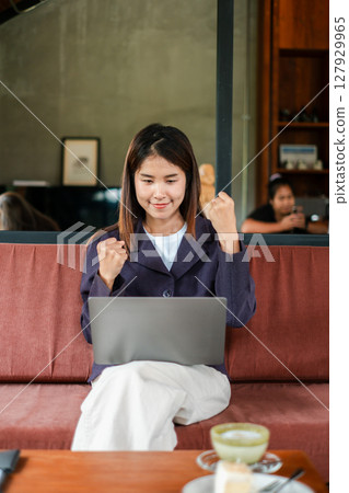 A joyful young woman celebrates a success while working on her laptop in a cozy cafe environment. A joyful young woman celebrates a success while working on her laptop in a cozy cafe environment. 127929965