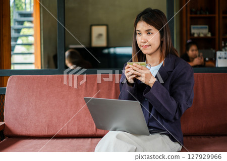 Woman enjoying a cup of coffee while working on a laptop in a comfortable living room environment. Woman enjoying a cup of coffee while working on a laptop in a comfortable living room environment. 127929966