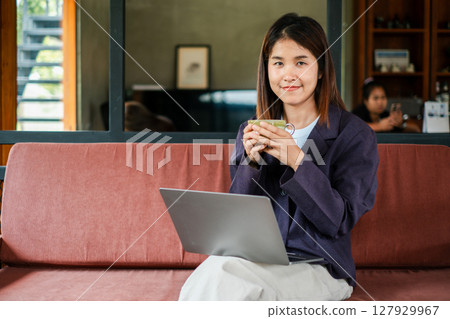 Smiling woman enjoying coffee while working on a laptop in a cozy, modern living room environment. Smiling woman enjoying coffee while working on a laptop in a cozy, modern living room environment. 127929967