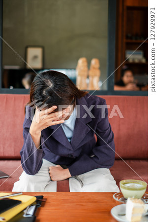 Woman in a cafe looking stressed, sitting with hand on forehead, reflecting in a cozy setting. Woman in a cafe looking stressed, sitting with hand on forehead, reflecting in a cozy setting. 127929971