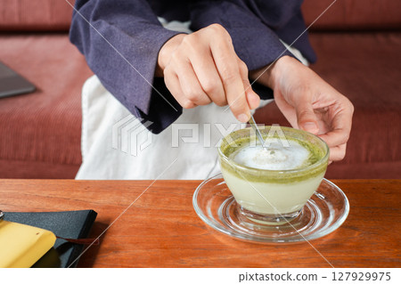 Close-up of hands stirring a frothy matcha latte in a glass cup, set on a wooden table with a cozy background. Close-up of hands stirring a frothy matcha latte in a glass cup, set on a wooden table with a cozy background. 127929975