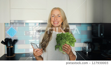 Cooking food, happy smiling mature woman with phone green lettuce vegetables in kitchen at home 127930003