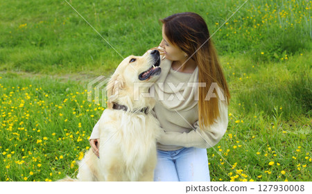 Happy owner young woman with Golden Retriever dog sitting on grass together in summer park 127930008