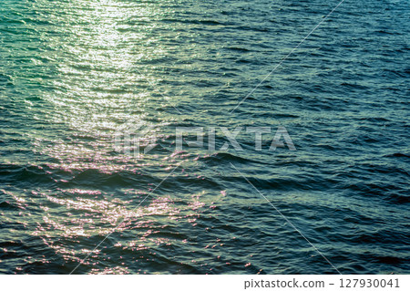 Golden sunlight reflects on rippling sea surface, casting sparkling trail over deep blue water. Concept of ocean, serenity, light playing across tranquil natural texture, background 127930041