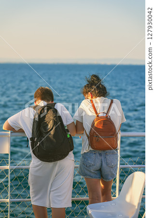 Young couple of friends, two travelers with backpacks lean on ferry railing, gazing at calm sea under warm evening light. Concept of travel, relaxation, shared adventure, tranquil maritime moments 127930042