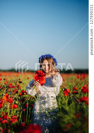 Young woman with blue flowers crown holding a bouquet of red poppies and gesturing in a field of papaver rhoeas 127930151