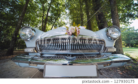 White vintage car adorned with a bouquet of flowers, parked in a wooded area, ready for a wedding ceremony 127930175