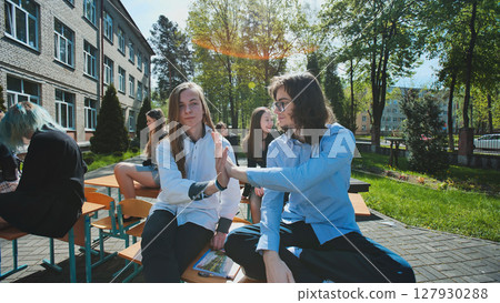Two high school male students giving high five sitting on school desks outdoors after class, celebrating their friendship 127930288