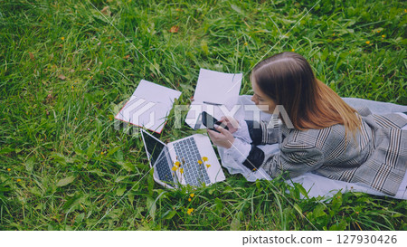 Young businesswoman lying on grass using laptop and smartphone, working remotely in a park surrounded by nature Young businesswoman lying on grass using laptop and smartphone, working remotely in a park surrounded by nature 127930426