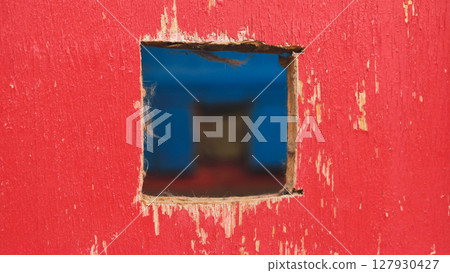 Square hole in red painted wood revealing blurred blue background 127930427
