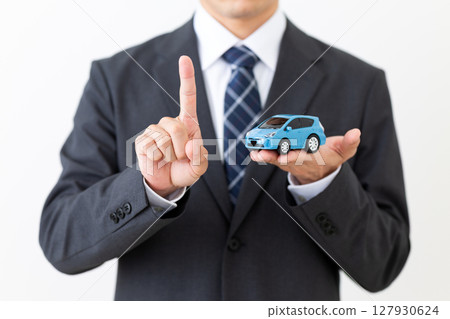 A middle-aged male businessman in a suit holding a model car and pointing his index finger 127930624