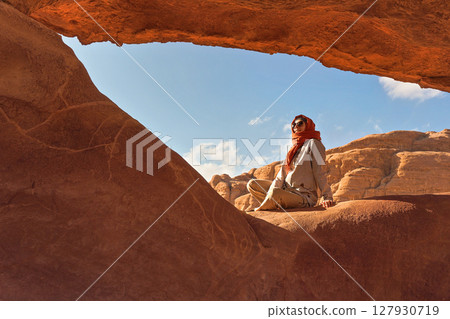 Young woman wearing warm jacket and scarf around head smiling, sitting at naturally formed rock window in wadi rum desert on sunny day 127930719
