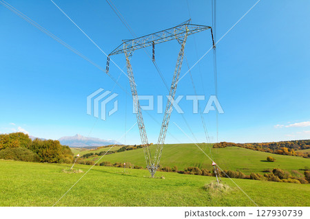 Large metal power pylon under electricity lines built in countryside with green grass covered hills and small forests, mount Krivan (Slovak symbol) at distance, blue sky background Large metal power pylon under electricity lines built in countryside with green grass covered hills and small forests, mount Krivan (Slovak symbol) at distance, blue sky background 127930739