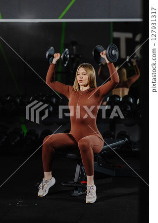 Determined sportswoman lifting dumbbells while sitting on bench in gym Determined sportswoman lifting dumbbells while sitting on bench in gym 127931317