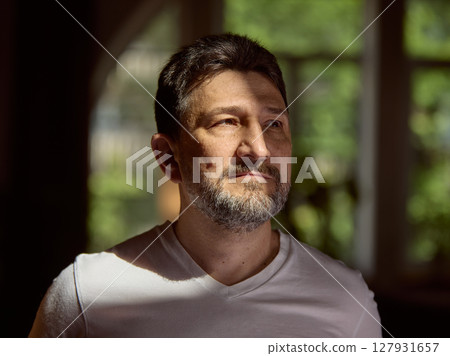 Emotional depth. Man in white shirt looking thoughtfully through sunlit window in cozy room 127931657