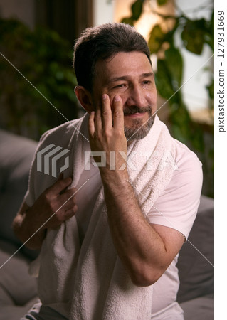 Hydration for men. Man in his 50s applying facial moisturizer at home with towel draped around neck 127931669