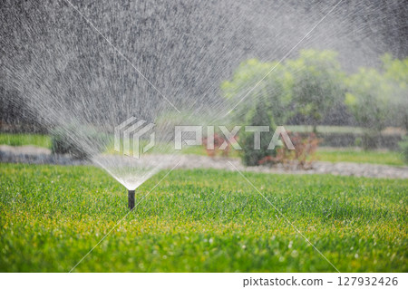 Garden sprinkler in action sprays water evenly across green lawn during summer irrigation in well-maintained residential backyard Garden sprinkler in action sprays water evenly across green lawn during summer irrigation in well-maintained residential backyard 127932426