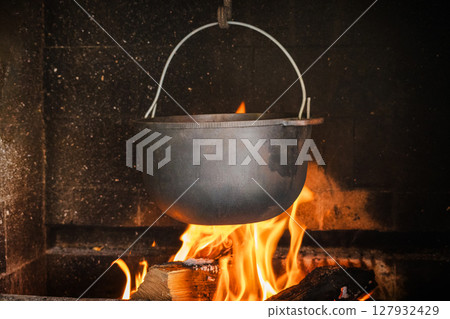 Iron cauldron hanging over burning firewood inside rustic fireplace Iron cauldron hanging over burning firewood inside rustic fireplace 127932429