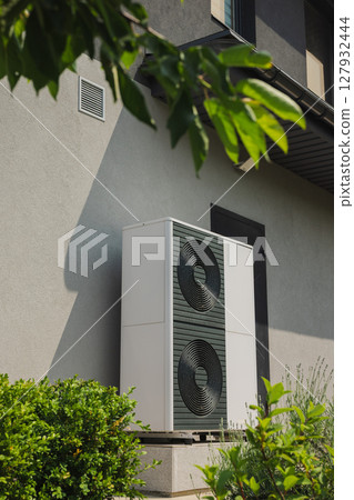 Vertical outdoor heat pump unit installed next to a house wall, surrounded by green shrubs in a modern landscaped garden 127932444
