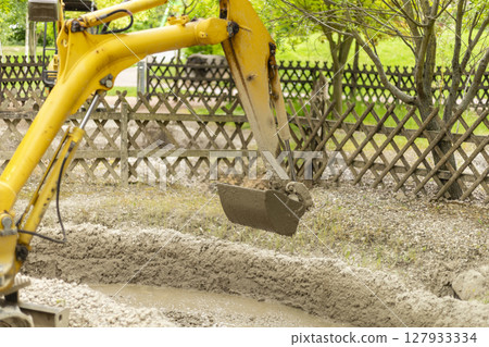 A yellow excavator performs earthmoving tasks in a construction zone surrounded by a wooden fence. The machinery lifts a bucket filled with soil under clear skies 127933334