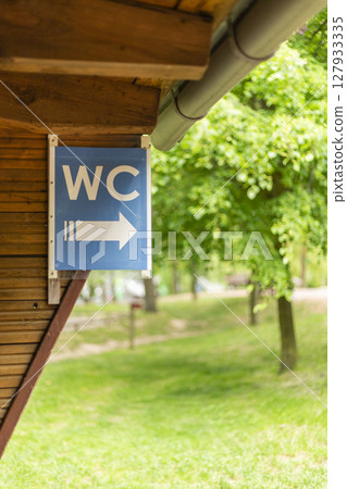 A sign indicating the location of the restroom is attached to a wooden structure, showing a clear arrow pointing to the right amidst lush greenery and trees in daylight 127933335