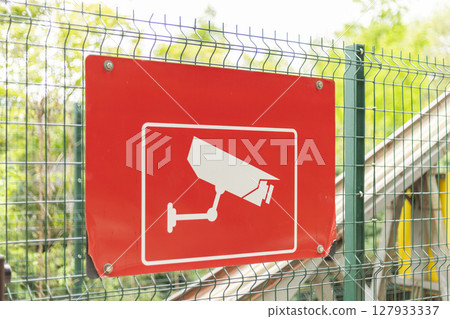 A bright red sign displaying a security camera graphic is mounted on a fence, alerting visitors to the presence of surveillance in the vicinity. The image captures greenery in the background A bright red sign displaying a security camera graphic is mounted on a fence, alerting visitors to the presence of surveillance in the vicinity. The image captures greenery in the background 127933337