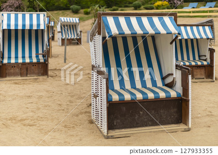 Blue and white striped beach chairs are arranged neatly on the sandy shore, creating a relaxing spot for visitors. The sun shines brightly, inviting leisure and enjoyment in this serene environment Blue and white striped beach chairs are arranged neatly on the sandy shore, creating a relaxing spot for visitors. The sun shines brightly, inviting leisure and enjoyment in this serene environment 127933355