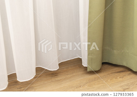 Gentle sunlight streams through a sheer white curtain alongside a solid green curtain, creating a warm, inviting atmosphere in a tranquil room with wooden floors 127933365