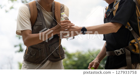 Father and Son Sharing Water on Hike. A father hands a water bottle to his son during their outdoor adventure. 127933758