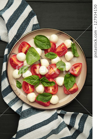 Italian Caprese salad with cherry tomatoes, mozzarella, basil on plate on black background. Vertical photo 127934150